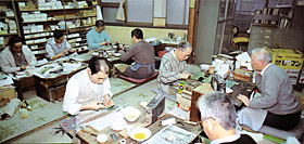 名村大成堂では、あぐら座で筆作りをする。作業台には、製筆の道具として、ガラス板、金櫛、半さし、手板、フノリや水を入れた容器などがみえる。
